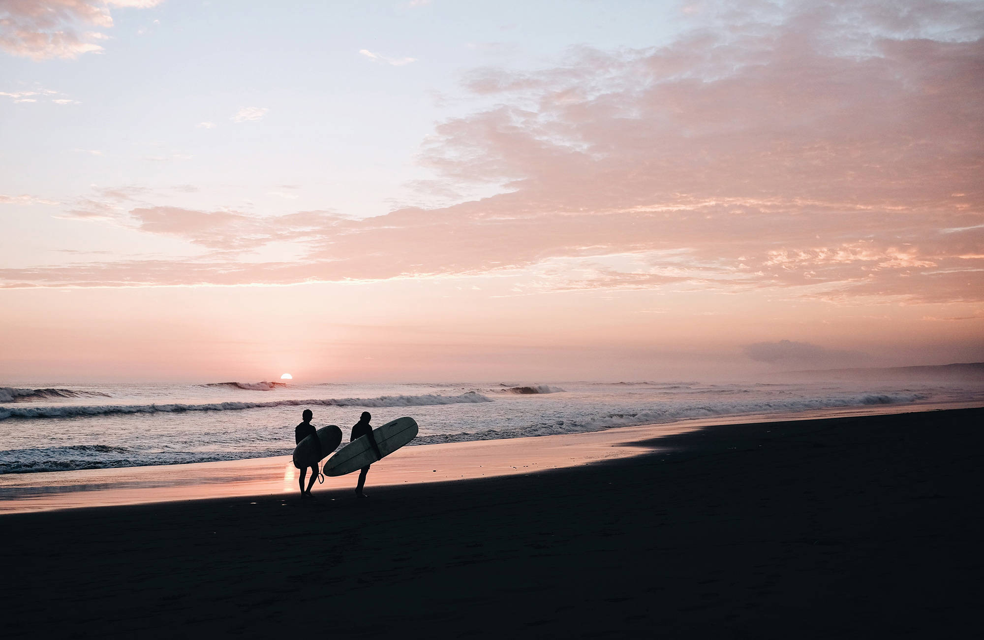 Två personer går på strand med surfbrädor i solnedgången.
