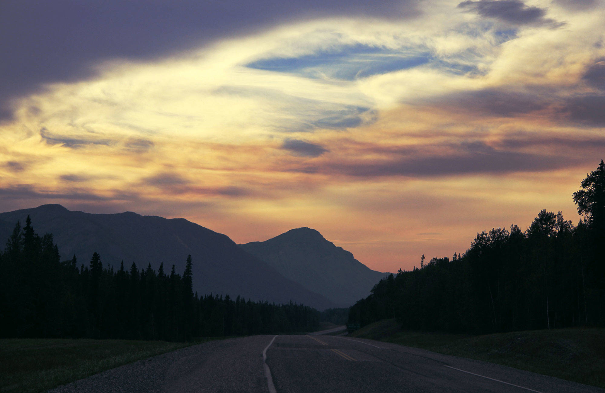 En väg omringad av berg och skog i solnedgången i Alaska i juni.