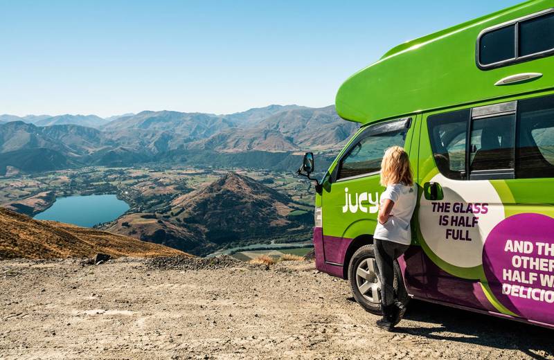 A girl leaning against a Jucy Condo campervan, looking out over a lake and mountains