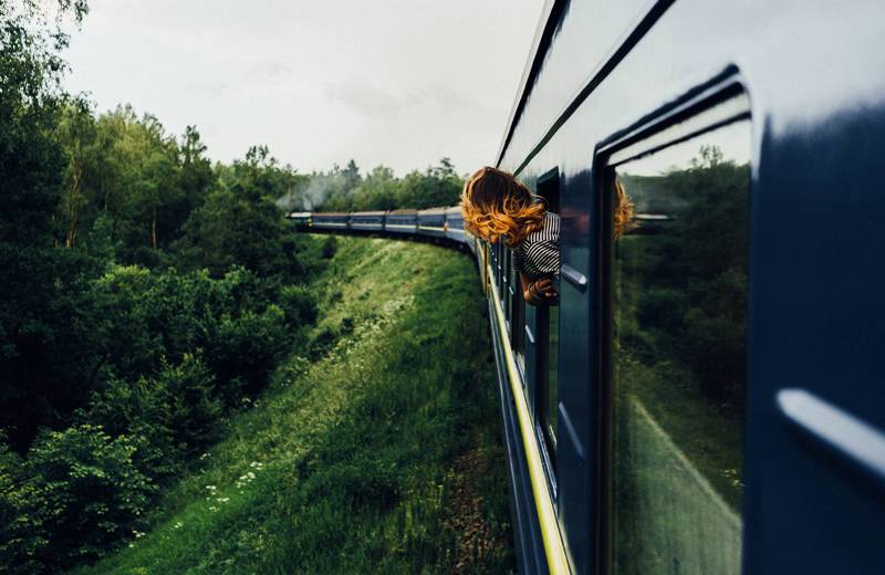Girl sticking her head out the window of a blue old-fashioned train that's in motion.