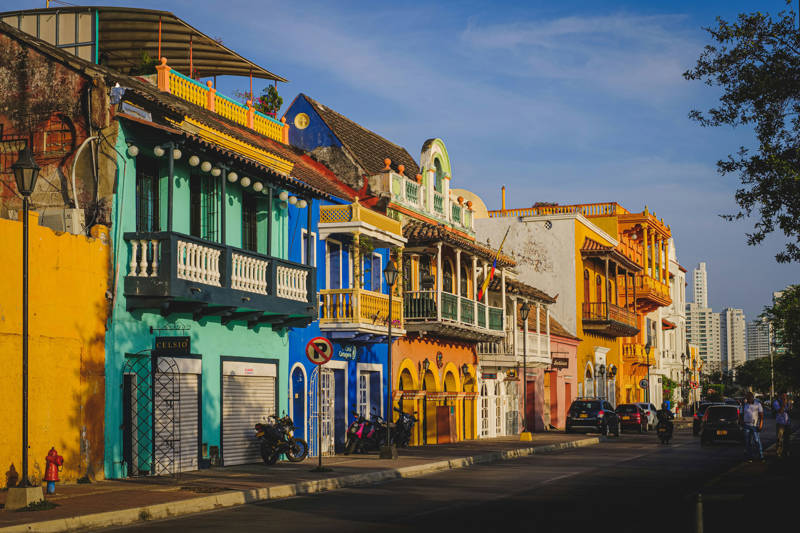 A street with in Cartagena with passing traffic and lots of colourful, colonial-style buildings in blue, teal, yellow and pink